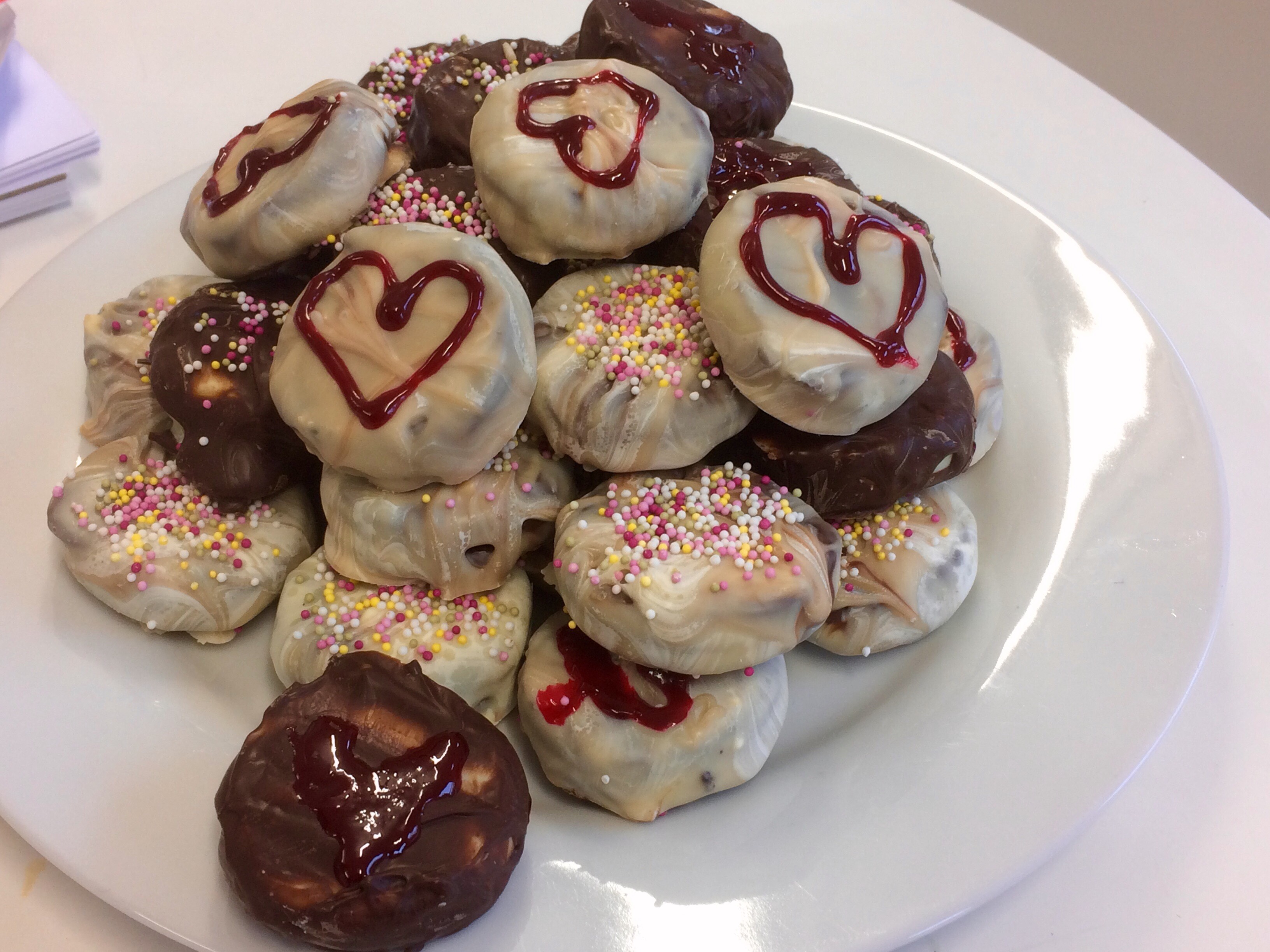 Double dipped oreos with milk chocolate and white chocolate, some have designs of hearts in icing on the top. The biscuits are arranged on a plate.
